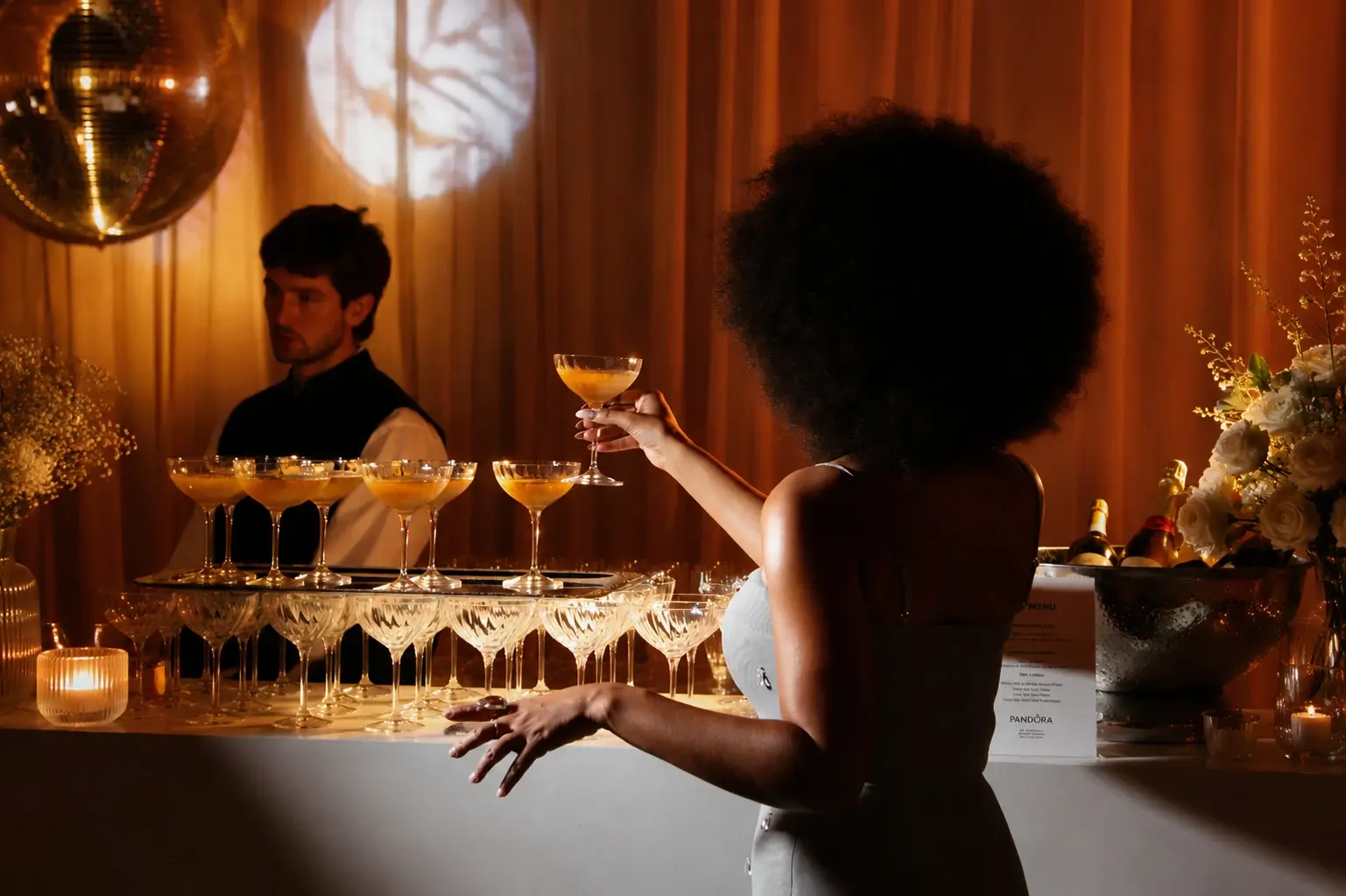 Une invitée saisit un cocktail sur un bar éclairé aux teintes ambrées